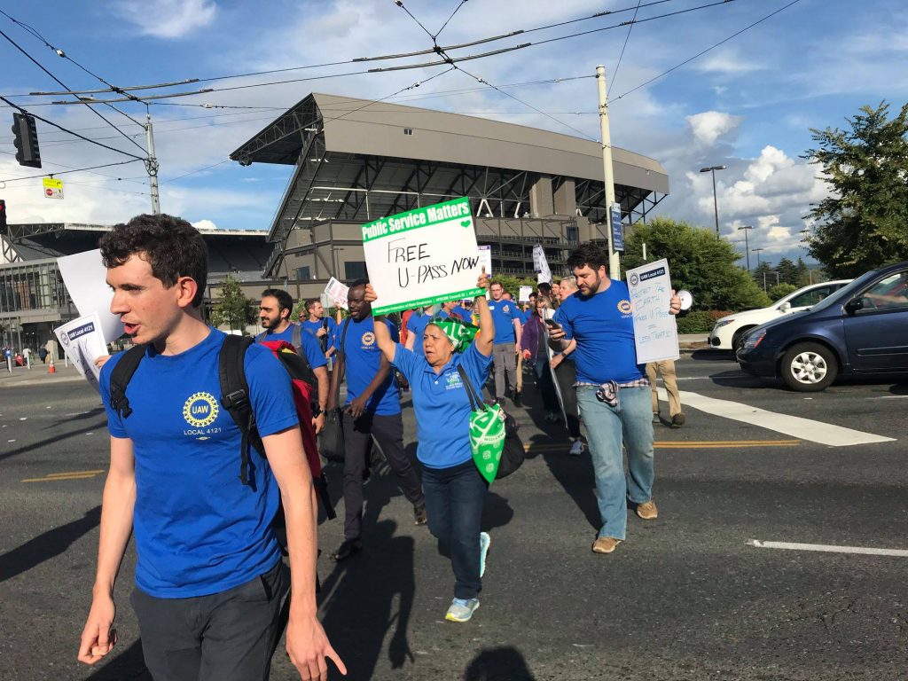 A crowd of people wearing blue UAW 4121 shirts crosses a street holding signs. One person in the center of the picture in a blue shirt holds a sign that reads "Free U-PASS Now"