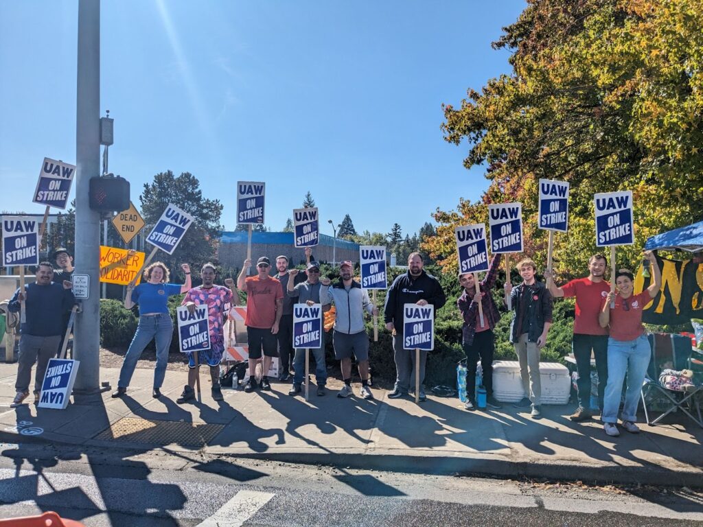 Approximately a dozen people stand on the picket line in front of a parts plant in Beaverton, Oregon holding signs that read “UAW on Strike” 