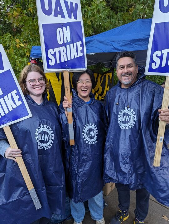 Alt text: three smiling UAW members wearing ponchos on Local 492’s picket line
