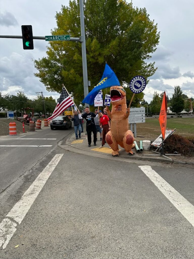 A person in an inflatable T-Rex costume waves the UAW flag in one hand and a UAW sign in another. Other people hold picket signs and another member waves the US flag